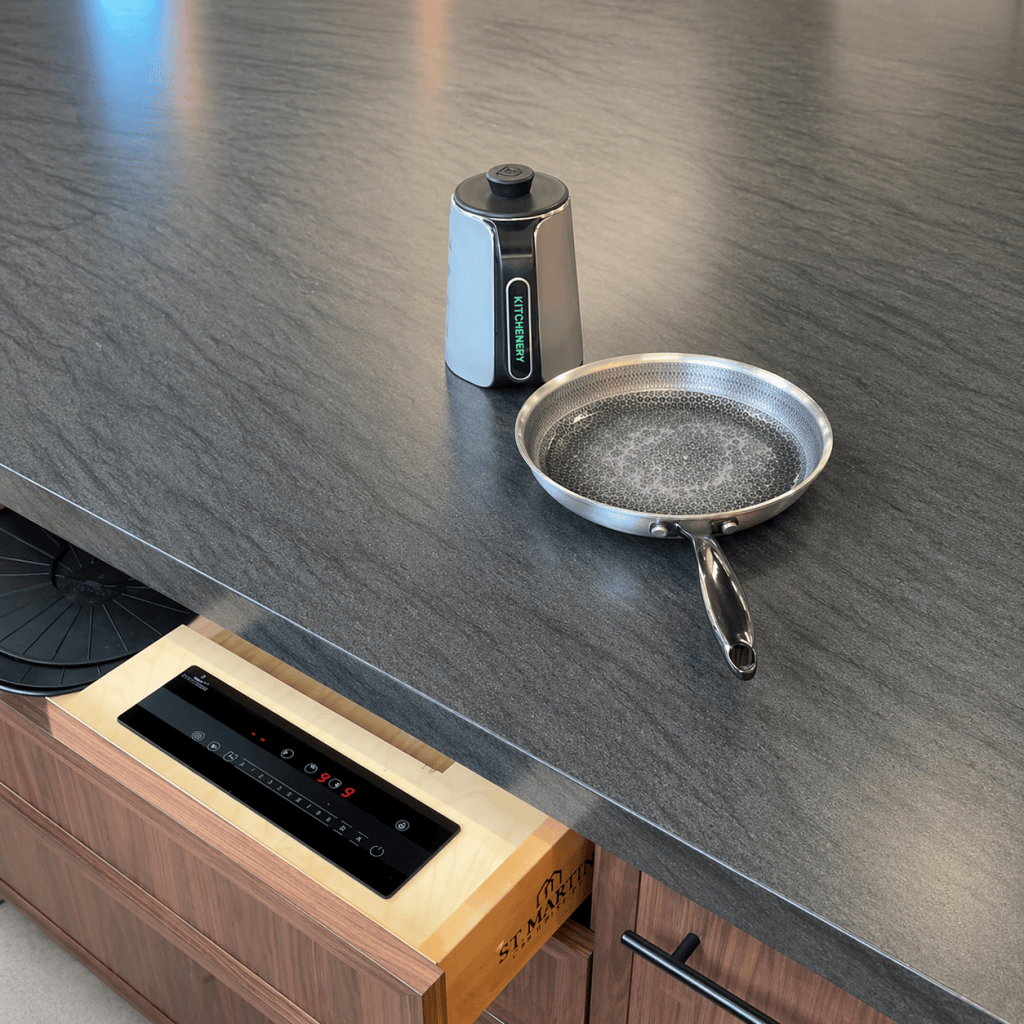 Stainless steel frying pan with a black handle on a dark gray kitchen counter beside a small vertical kitchen gadget.